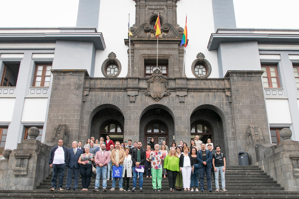 Foto de grupo de las personas asistentes al acto, bajo la bandera arco iris ondeando en la fachada del Edificio Central.